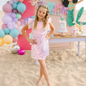 Young girl in a sequin dress standing in front of a colorful balloon arch and mermaid-themed decorations.