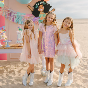 Three young girls in dresses standing in front of a colorful balloon arch and party setup.