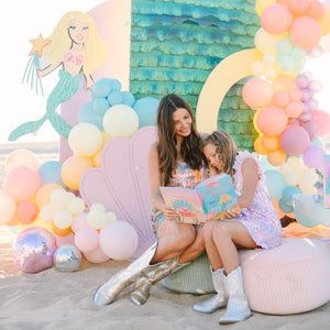 A mom and a daughter sitting under a colorful balloon arch with a mermaid-themed backdrop.