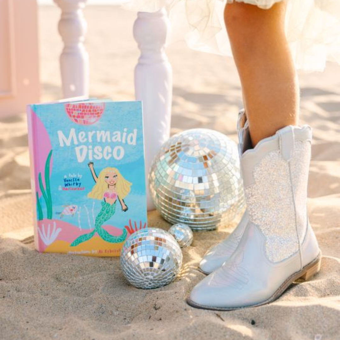Book titled 'Mermaid Disco' on sand with disco balls and silver Disco Dreams Wild + Rae boots.