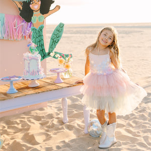 Young girl in a pink dress standing on a sandy beach with a mermaid-themed cake and decorations.