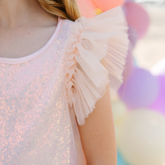 Close-up of a person wearing a light pink sequin top with ruffled sleeves against a colorful blurred background.