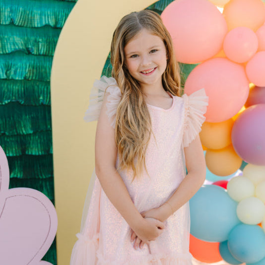 Young girl in a pink dress standing in front of colorful balloons and a decorative arch.