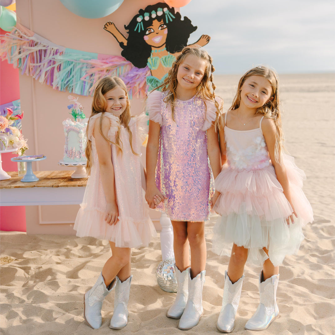 Three young girls in dresses standing in front of a colorful balloon arch and party setup.