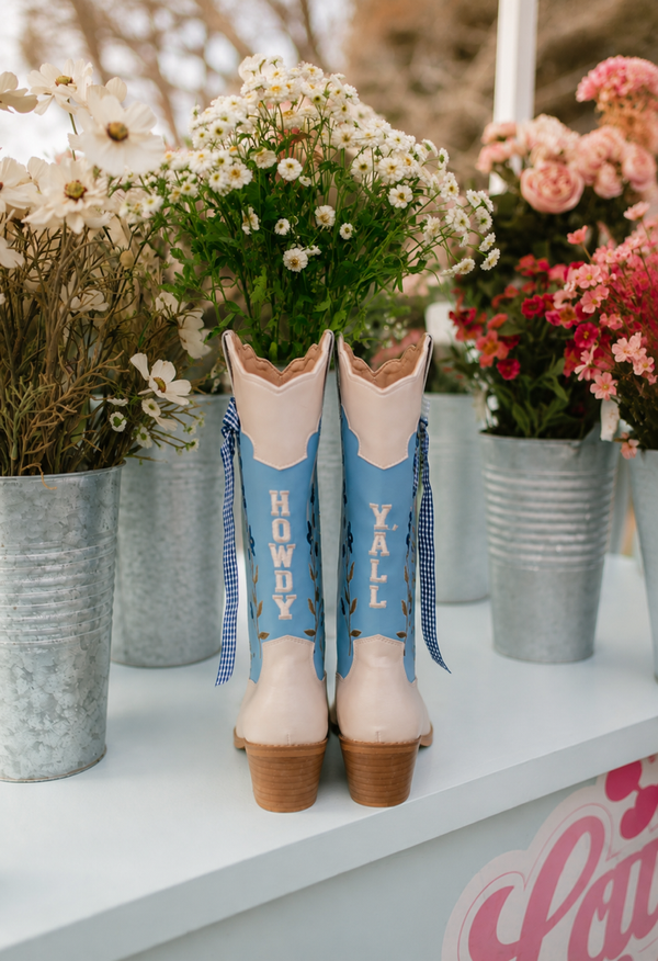 Decorative cowboy boots with 'Howdy' and 'Y'all' text, surrounded by potted plants on a shelf.
